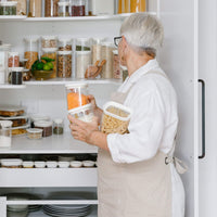 Person organising Tallow Rectangle glass pantry containers on open kitchen shelving, showing the jars in multiple shelf heights.