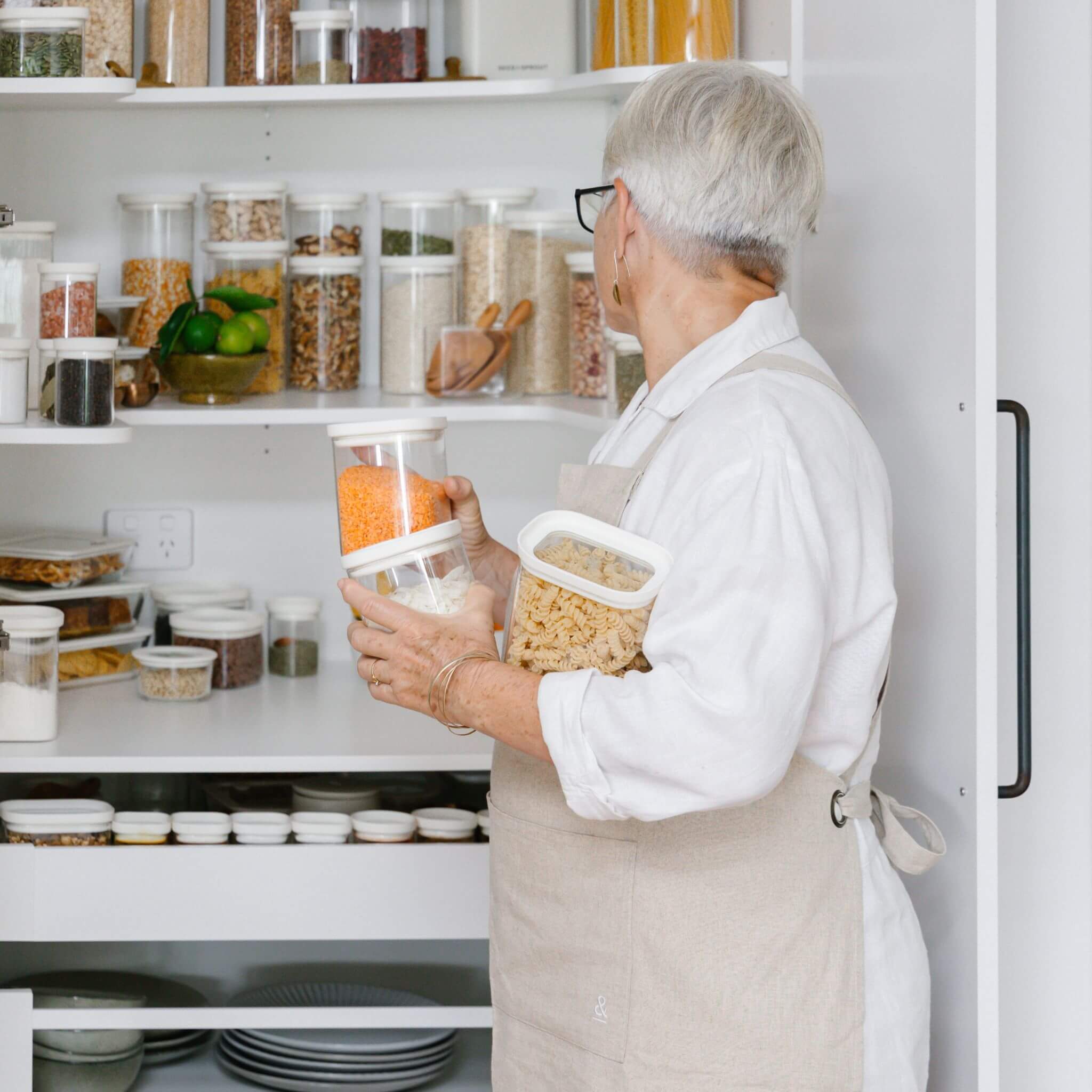 Person organising Tallow Rectangle glass pantry containers on open kitchen shelving, showing the jars in multiple shelf heights.
