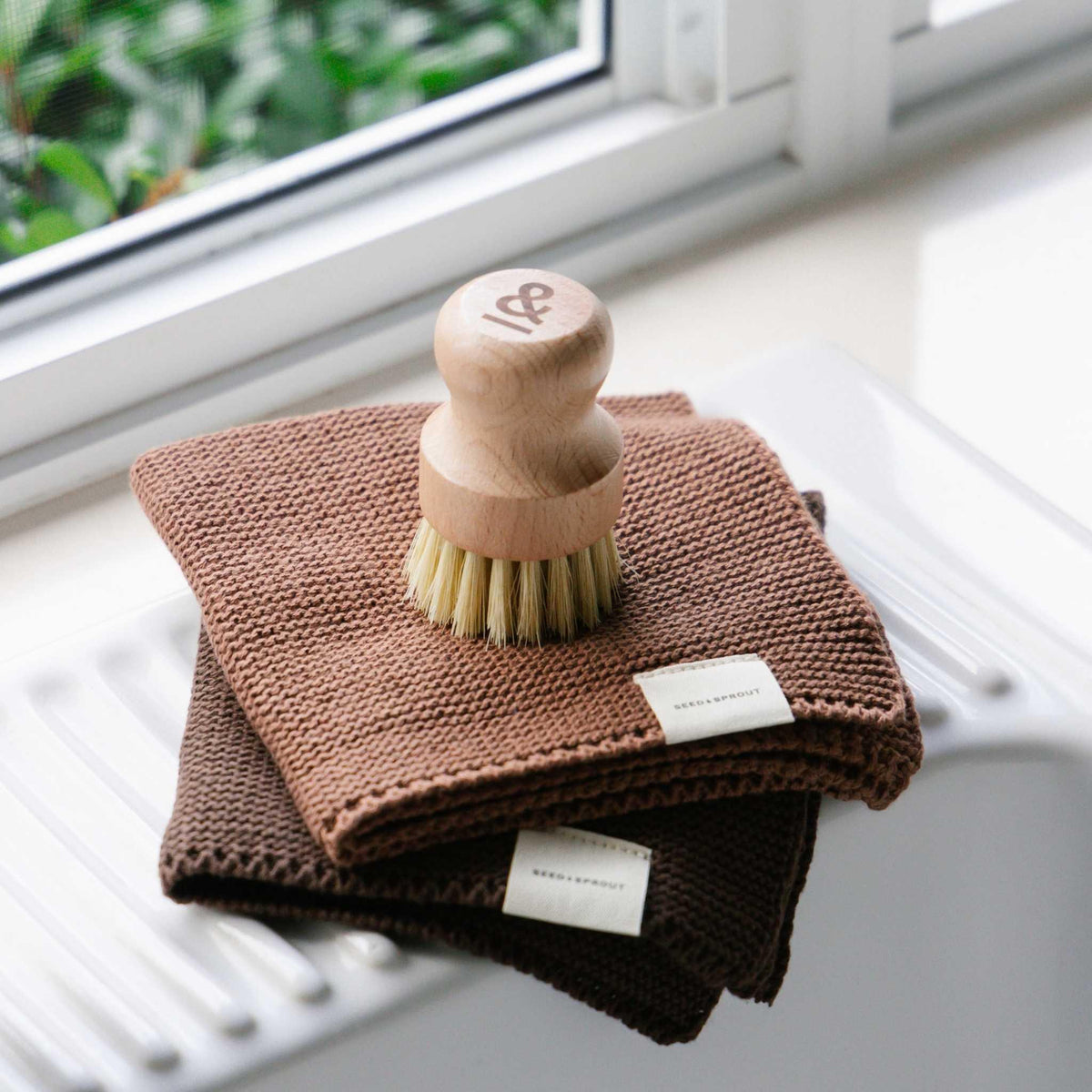 Cotton Knitted Dishcloths set of 2 in Cinnamon & Nutmeg, folded on a sink drainboard with a wooden pot scrubber on top, showing the knit texture and brand labels