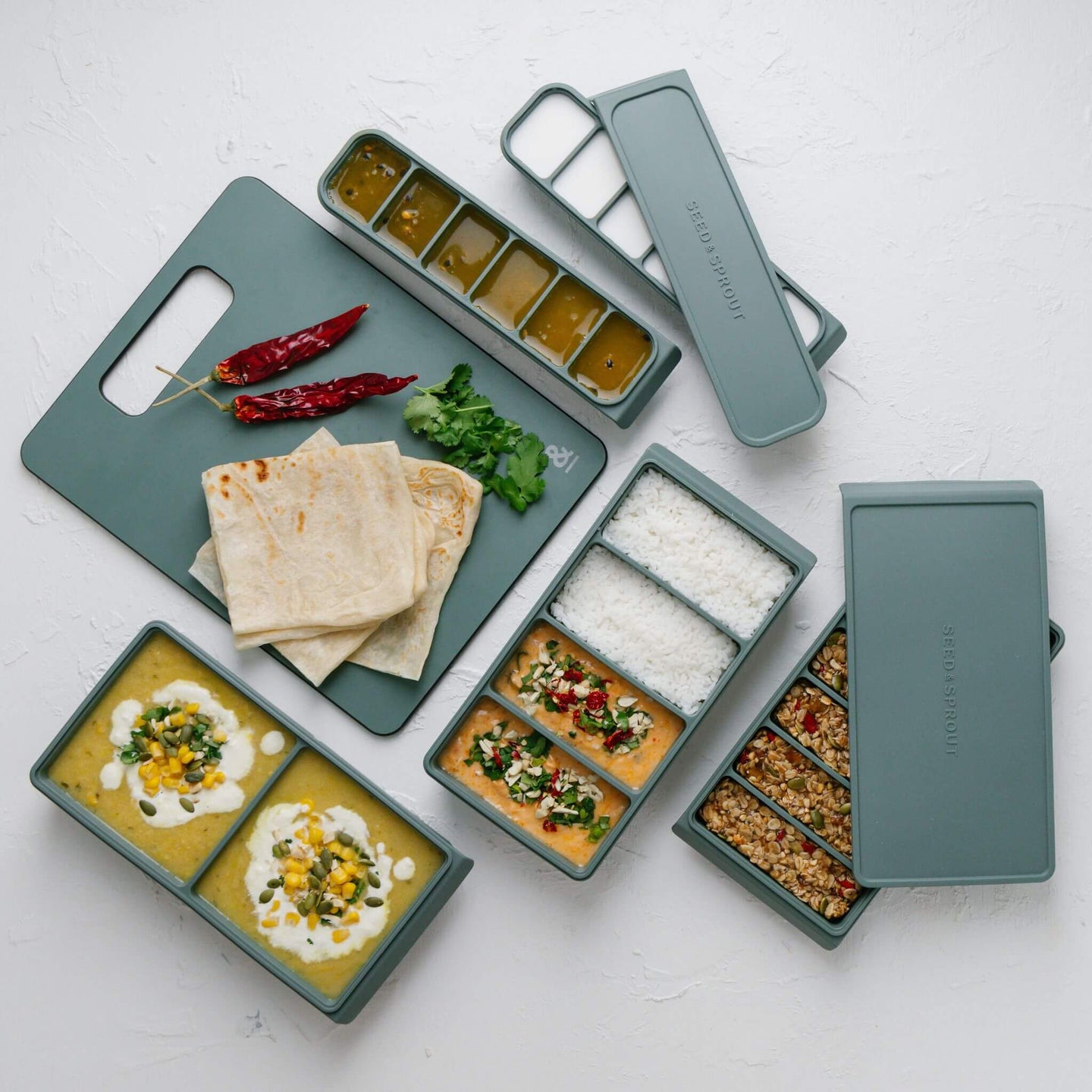 Freezer Mould Set in sage green displayed overhead with prepped meals — curry, rice, granola bars, and sauce portions — alongside a matching silicone cutting board with flatbreads, chillies, and fresh herbs