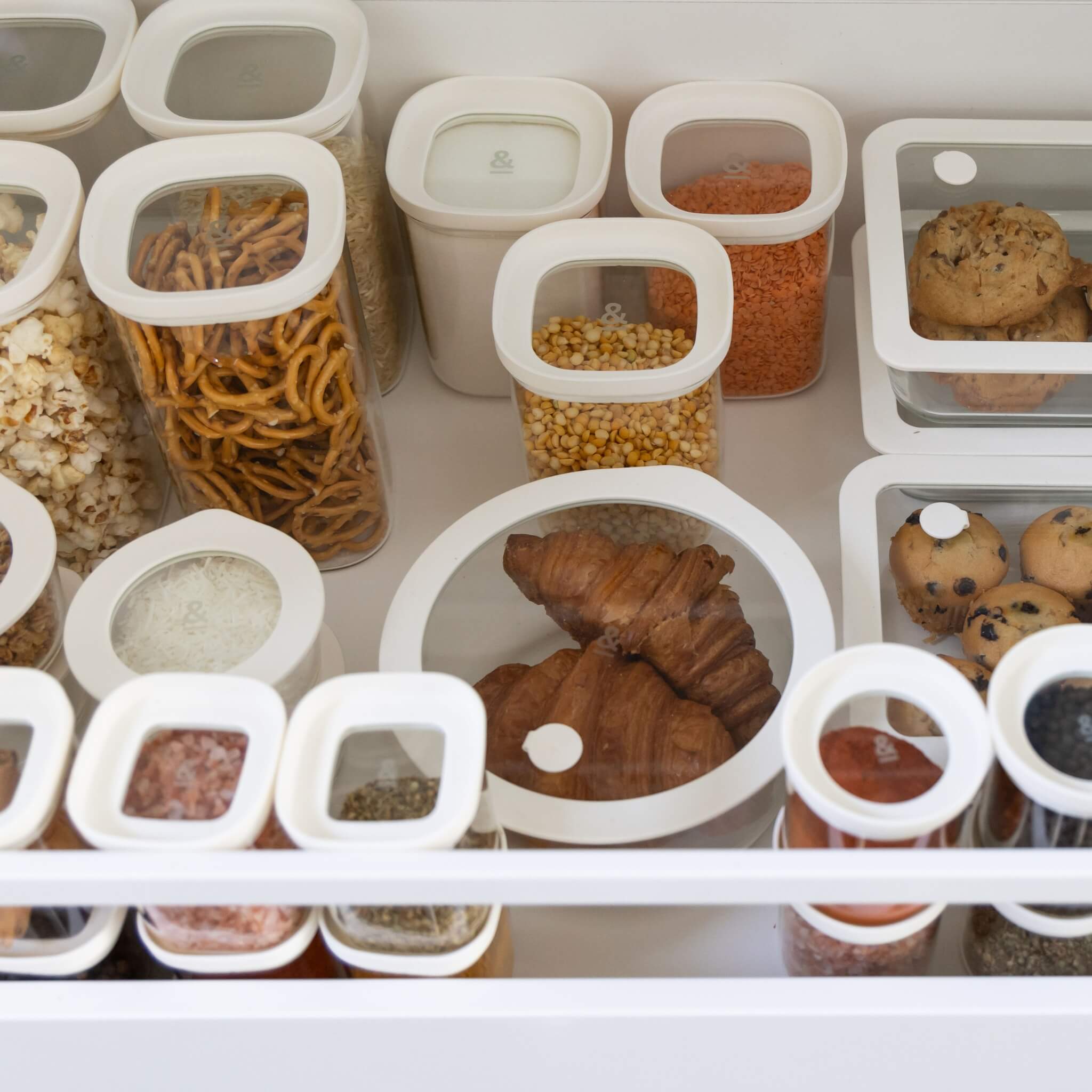 A pantry drawer with different glass food containers that have white lids. The containers contain breads, grains, snacks and condiments